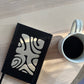 White mug with black coffee on a wooden surface next to a black notebook with white patterns
